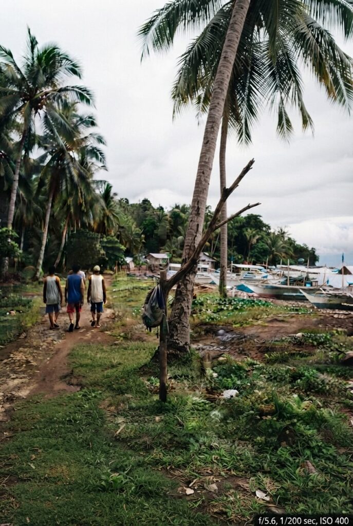 Three people walk away down a dirt path lined with palm trees in a village setting. Several boats are beached on the right, and a backpack hangs from a leaning palm trunk.