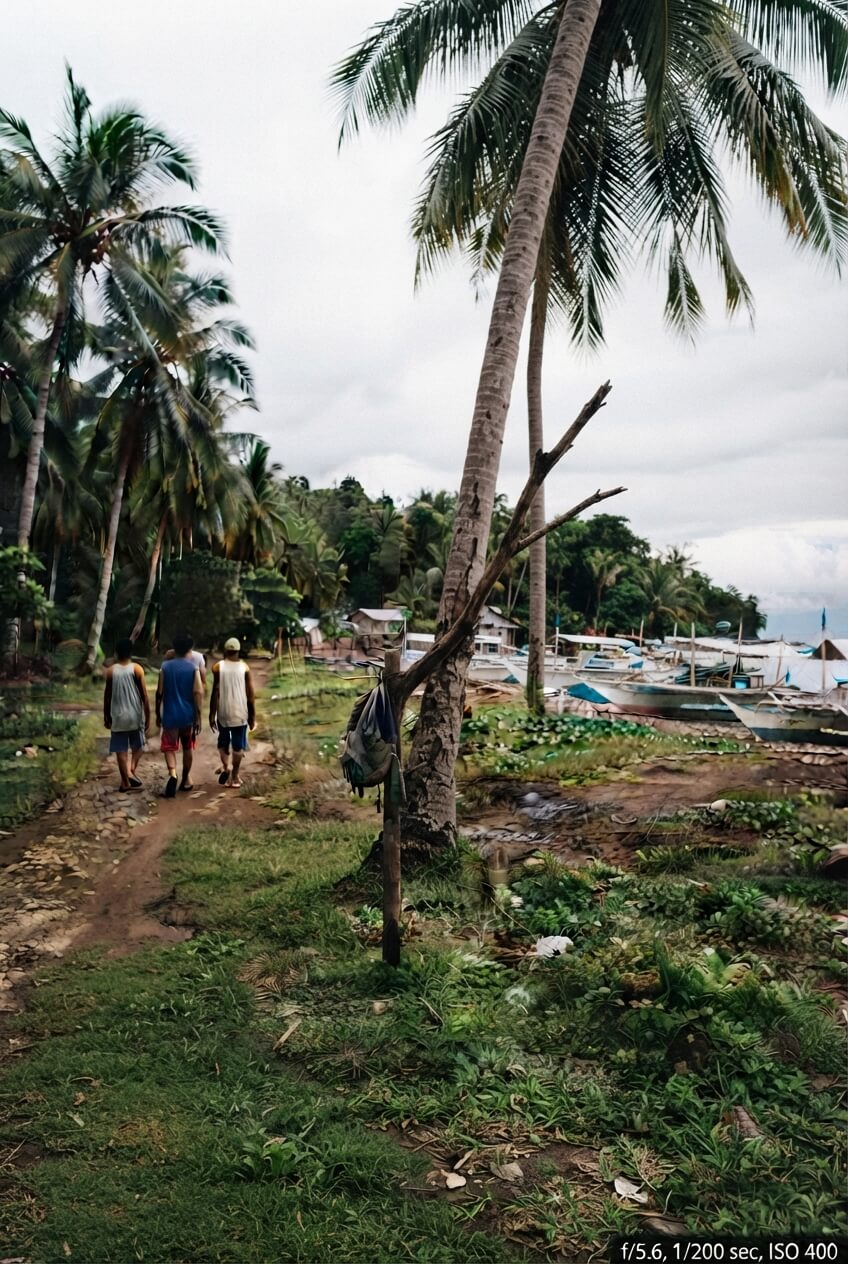 Three people walk away down a dirt path lined with palm trees in a village setting. Several boats are beached on the right, and a backpack hangs from a leaning palm trunk.